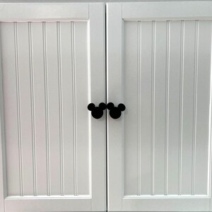 May include: A pair of white cabinet doors featuring vertical beadboard paneling and two black cabinet knobs shaped like the silhouette of a mouse head. The doors are positioned side by side with the knobs meeting in the center of the frame.