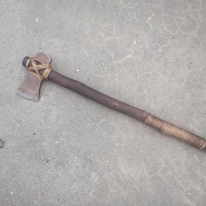 Könnte beinhalten: Eine rustikale Axt mit einem dunkelbraunen Holzgriff und einer Metallklinge. Die Klinge ist mit Bindfaden am Griff befestigt. Der Griff ist in der Nähe der Klinge und am Ende mit Bindfaden umwickelt.