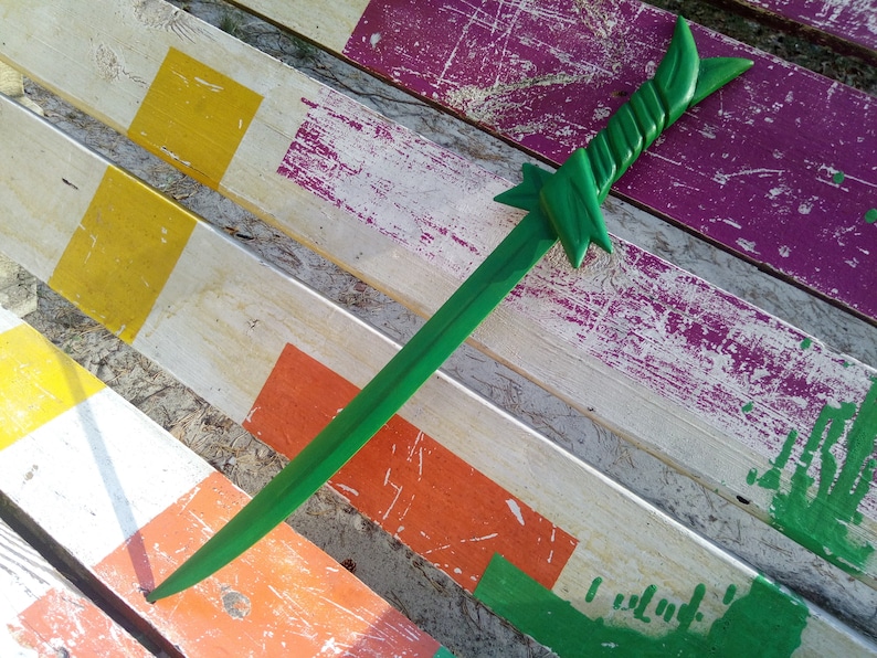 May include: A green wooden sword with a twisted handle lies on a weathered wooden surface painted with stripes of yellow, orange, green, and purple.