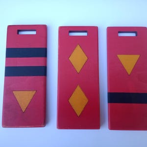 Puede incluir: Tres placas rectangulares de madera roja con diseños geométricos. Una tiene rayas negras y un triángulo naranja, otra tiene dos rombos naranjas y la última tiene un triángulo naranja y una franja negra.