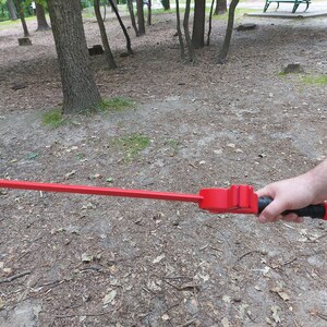 May include: A red, long, straight, metal sword with a black handle. The sword is held by a person's hand in a park setting.