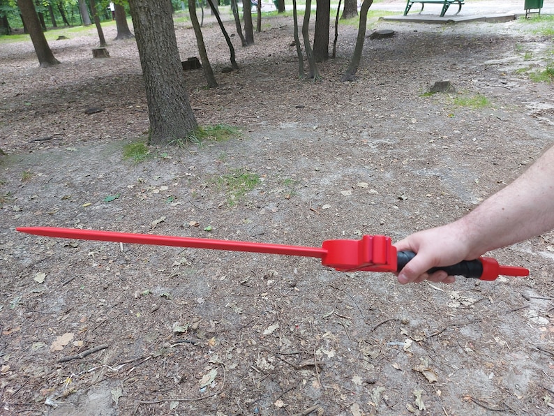 May include: A red, long, straight, metal sword with a black handle. The sword is held by a person's hand in a park setting.