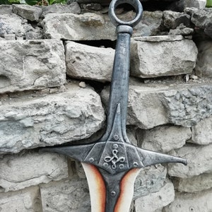 May include: A silver sword with a decorative handle and a red and white blade. The sword is leaning against a stone wall.
