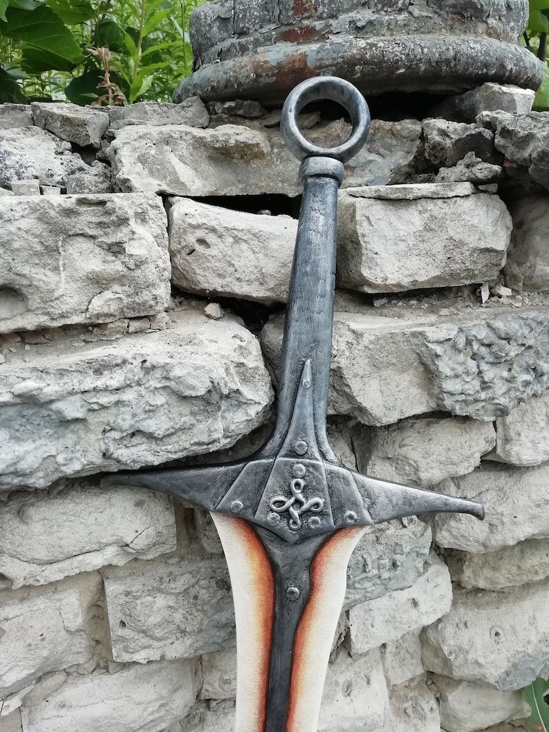 May include: A silver sword with a decorative handle and a red and white blade. The sword is leaning against a stone wall.