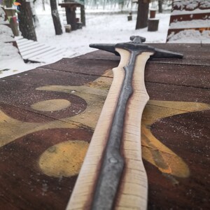 May include: A wooden sword with a silver blade, lying on a wooden surface with a brown and yellow paint design.