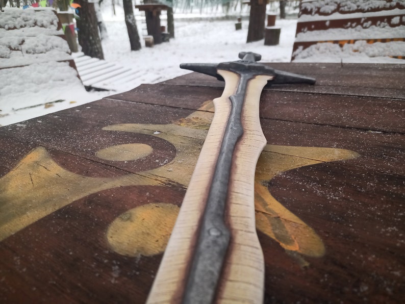 May include: A wooden sword with a silver blade, lying on a wooden surface with a brown and yellow paint design.