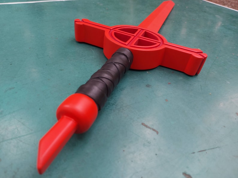 May include: A red wooden sword with a black grip and a cross-shaped design on the blade.