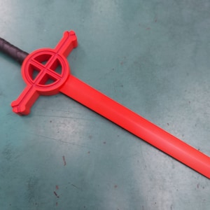 May include: A red plastic sword with a black handle and a circular design on the guard. The design features a cross inside a circle.