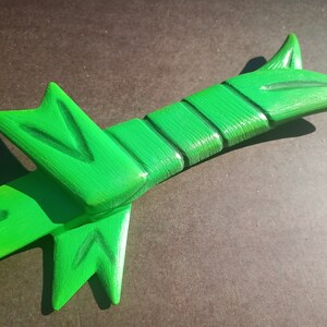May include: A green wooden toy sword with a textured surface and a pointed tip. The sword has a handle with a decorative design.