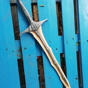 May include: A silver and brown sword with a decorative handle and a pointed blade. The sword is lying on a blue wooden surface.