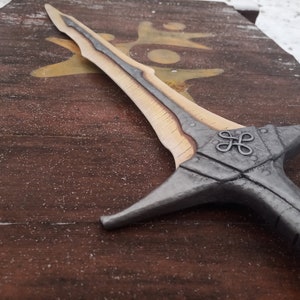 May include: A silver and wood sword with a decorative knot design on the hilt. The sword is lying on a wooden surface.