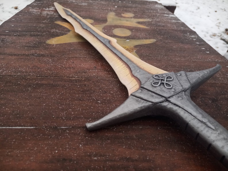 May include: A silver and wood sword with a decorative knot design on the hilt. The sword is lying on a wooden surface.