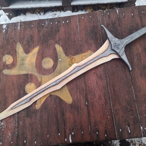 May include: A silver and gold sword with a decorative handle and a ring on the pommel. The sword is lying on a wooden surface.