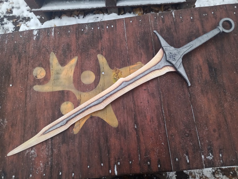 May include: A silver and gold sword with a decorative handle and a ring on the pommel. The sword is lying on a wooden surface.