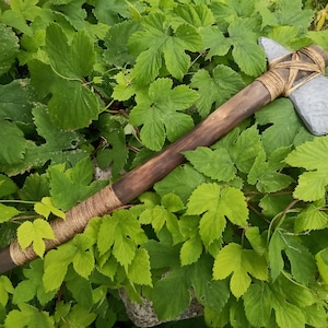 May include: A handcrafted axe with a wooden handle and a gray stone head, wrapped with natural twine. The axe is set against a backdrop of lush green leaves, showcasing its rustic design. The axe measures approximately 24 inches in length.