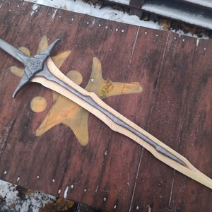 May include: A silver and wood sword with a decorative handle and a black and silver blade. The sword is lying on a wooden surface.