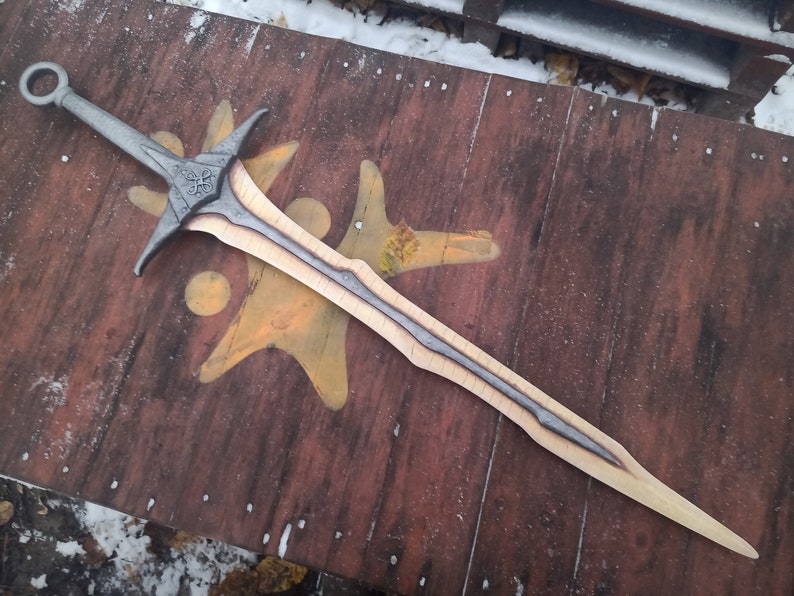 May include: A silver and wood sword with a decorative handle and a black and silver blade. The sword is lying on a wooden surface.