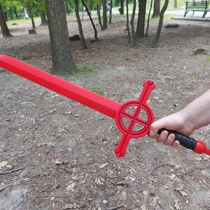 May include: A red sword with a black handle and a circular design on the guard. The sword is held by a person's hand in a forest setting.
