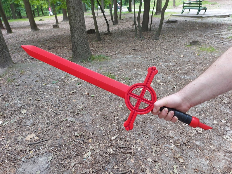 May include: A red sword with a black handle and a circular design on the guard. The sword is held by a person's hand in a forest setting.