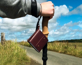 May include: A brown leather wallet attached to a hiking stick with a black strap. The stick has a wooden handle and a black metal shaft.