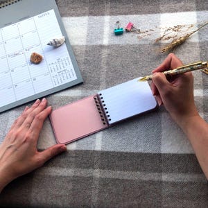 May include: A small, pink-covered notepad with lined paper is open, ready for writing, with a pen held above it. A calendar, seashells, and paper clips are also visible, suggesting a workspace or planning theme.