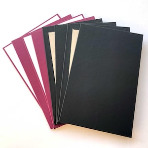May include: A selection of coloured cardstock fanned out. The cardstock includes black, white, and burgundy shades. The cardstock is arranged in a fan shape, with the black cards at the front. The paper is rectangular and has a smooth surface.