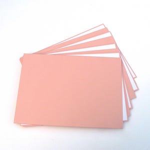 May include: A collection of pale pink rectangular cards fanned out against a white backdrop. The cards have a smooth, matte finish, with white edges visible between each card. The arrangement suggests stationery or paper products.
