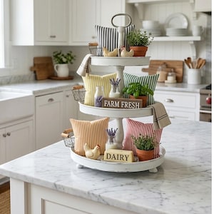 May include: A three-tiered white serving tray with decorative pillows, plants, and small baskets. The pillows are striped in various colors, including yellow, orange, red, and blue. The tray is decorated with small chickens and a sign that reads "FARM FRESH" and "DAIRY".