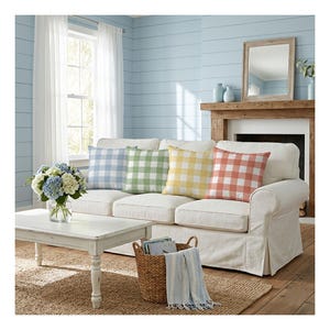 May include: A set of four square throw pillows featuring a classic gingham check pattern in light blue, sage green, soft yellow, and coral orange, arranged neatly on a white slipcovered sofa with a matching white coffee table in front.