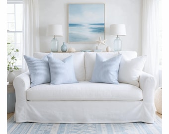 May include: A white slipcovered sofa featuring two solid white throw pillows and two light blue and white striped throw pillows. The sofa is positioned in a room with a light blue and white patterned area rug and white curtains.
