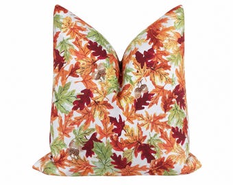May include: A square throw pillow featuring a white background with a dense, colorful pattern of autumn leaves in shades of orange, red, yellow, and green, interspersed with small brown acorns throughout the entire surface of the fabric.