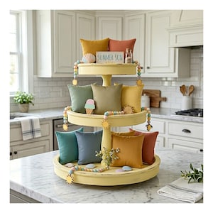May include: A three-tiered, yellow decorative tray filled with colorful throw pillows in shades of teal, blue, green, gold, and coral. The top tier has a sign that reads "SUMMER SUN". Decorative beads and wooden sun accents add to the summer theme.