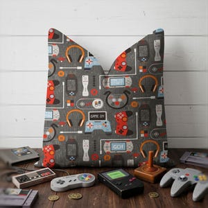 May include: Gray throw pillow with a pattern of video game controllers, headphones, and game consoles in red, blue, orange, and white. The pillow is surrounded by vintage game consoles and controllers on a wooden surface.