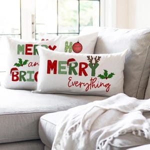 May include: White decorative throw pillows with Christmas-themed text, including "Merry and Bright" and "Merry Everything." The text is in red and green, with festive accents like Santa hats, holly, and reindeer antlers. The pillows are arranged on a gray sofa.