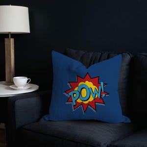 Puede incluir: Cojín azul con un gráfico POW! de estilo cómic rojo, amarillo y azul.