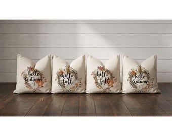 May include: Four square throw pillows with a light beige fabric base. Each pillow features a central circular wreath design composed of autumn leaves, small pumpkins, and the black cursive text "hello autumn" or "hello fall" printed in the center.