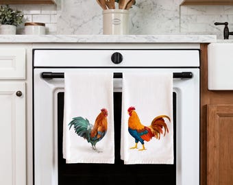 May include: Two white kitchen towels hanging on an oven handle. The left towel features a rooster with orange, brown, and dark green feathers. The right towel displays a rooster with bright orange, red, and blue plumage against a plain white background.