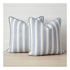 May include: Two square throw pillows featuring a vertical striped pattern in shades of light blue and off-white. Each pillow is finished with a dark blue piped edge along the seams, creating a defined border around the striped fabric design.