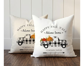 May include: Two decorative throw pillows with fall-themed designs. One pillow is off-white, the other is white. Each features a black and white checkered truck carrying pumpkins, with text that reads "Happy Fall Y'all" and "Pumpkin Patch".