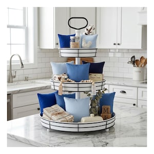 May include: A three-tiered serving tray with a white frame and black accents, filled with blue pillows, decorative items, and kitchen accessories. The top tier holds a jar labeled "FRESH EGGS". The tray is on a marble countertop.