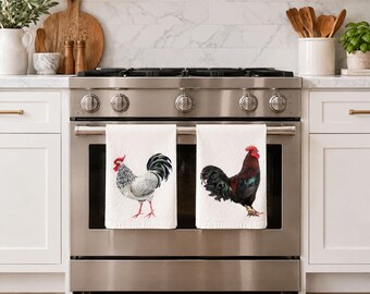 May include: Two white kitchen towels hanging on a stainless steel oven handle. The left towel features a light gray and white rooster with a red comb. The right towel features a dark brown and black rooster with a red comb and yellow leg band.