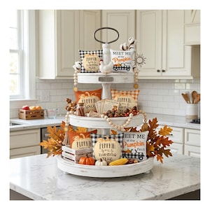May include: A three-tiered white serving tray decorated for fall. The tray features small pillows with pumpkin and plaid patterns, pumpkins, leaves, and the text "Meet Me at the Pumpkin Patch". The tray is on a white countertop.