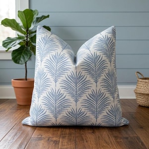 May include: A decorative pillow with a white background and a repeating pattern of blue palm fronds. The pillow is placed on a wooden floor, with a potted plant and a woven basket in the background. The pillow has a soft, inviting appearance.