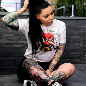 May include: A woman with long black hair pulled back in a ponytail is wearing a white t-shirt with a red heart and the text "True Love" printed on it. She has several tattoos on her arms and legs, including a black and white dog with pink flowers on her left leg. She is wearing black and white sneakers.
