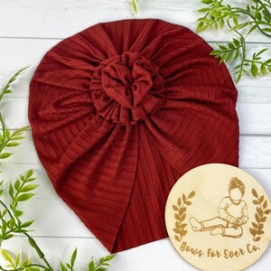 Peut inclure: Un turban en côtes de couleur bordeaux avec un gros nœud sur le dessus. Le turban est posé sur une surface en bois blanc avec une pancarte ronde en bois sur laquelle est inscrit "Bows for Ever Co."