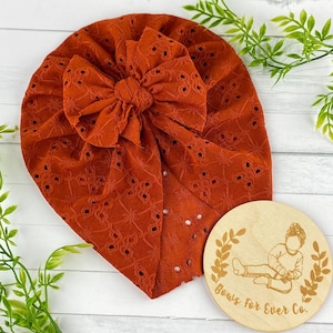 May include: A rust-coloured turban with a large bow and eyelet detailing. The turban is made of a soft, stretchy fabric and is perfect for adding a touch of style to any outfit. The image also includes a wooden sign with the text "Bows for Ever Co."