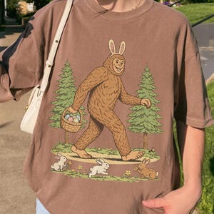 May include: Brown t-shirt featuring a Bigfoot character wearing bunny ears and carrying a basket of Easter eggs. The design includes green trees and three rabbits. The shirt is a relaxed fit.