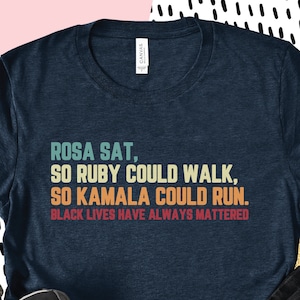 May include: A navy blue t-shirt with the text "ROSA SAT, SO RUBY COULD WALK, SO KAMALA COULD RUN. BLACK LIVES HAVE ALWAYS MATTERED" printed in a retro style.