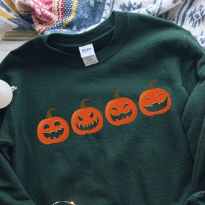 K&ouml;nnte beinhalten: Dunkelgr&uuml;nes Sweatshirt mit vier orangefarbenen K&uuml;rbissen mit unterschiedlichen Gesichtern. Die K&uuml;rbisse sind horizontal angeordnet. Wei&szlig;e Turnschuhe und Weihnachtsdekorationen sind im Hintergrund zu sehen.