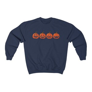 K&ouml;nnte beinhalten: Marineblaues Sweatshirt mit vier orangefarbenen K&uuml;rbisgesichtern mit unterschiedlichen Schnitzereien. Die K&uuml;rbisse sind horizontal angeordnet. Ein lang&auml;rmliges Oberteil f&uuml;r Halloween.